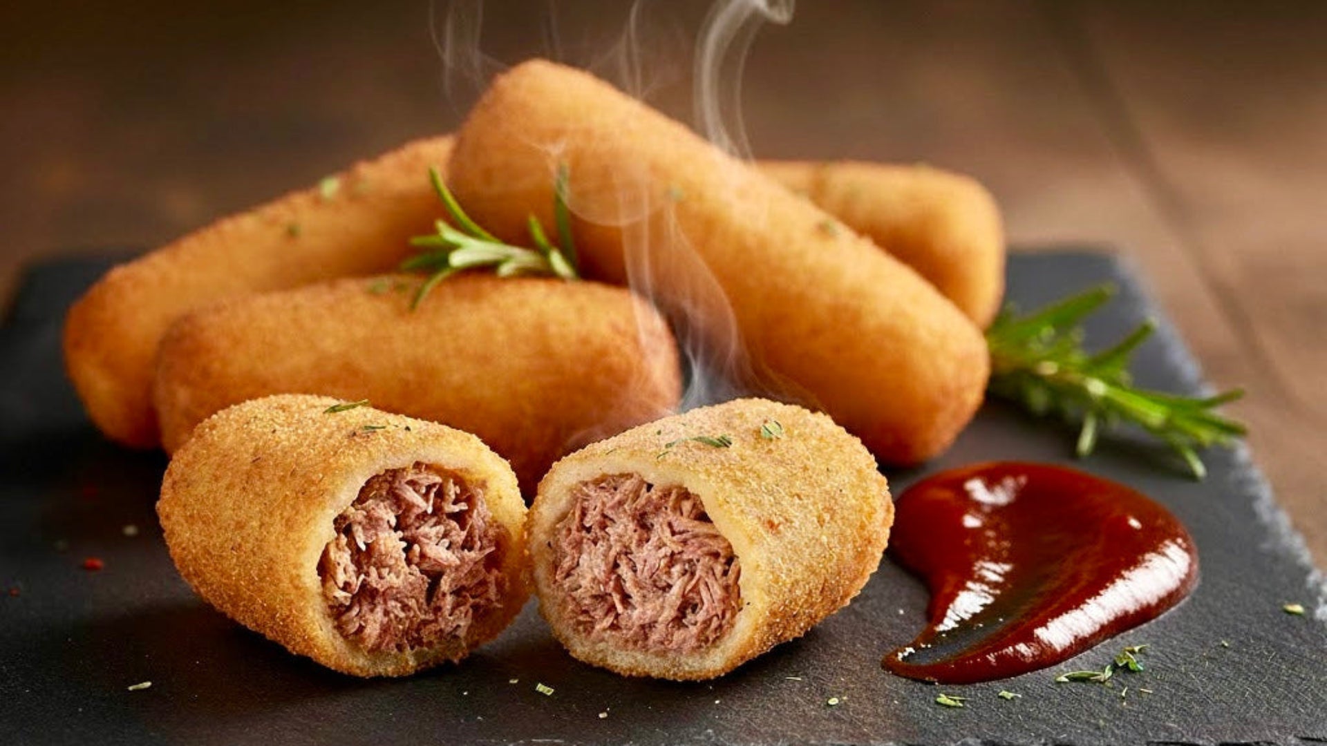 Fried pulled pork croquettes with a side of bbq sauce on a dark slate board with steam rising.